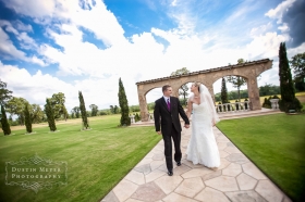 A bride and groom hold hands and smile at each other while walking down the aisle together in front of a stone arch with grass and trees and blue sky behind them for their wedding day portraits.
